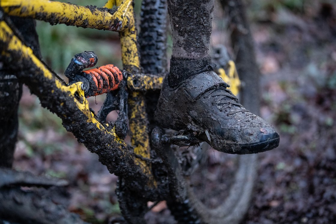 A close-up of a mountain bike heavily covered in mud, along with a similarly dirty cycling shoe. The yellow bike frame and suspension are visible, while the ground appears wet and covered with leaves.