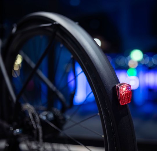 Gros plan d’un feu arrière de vélo fixé au garde-boue et allumé ; lumières urbaines floues en arrière-plan nocturne.