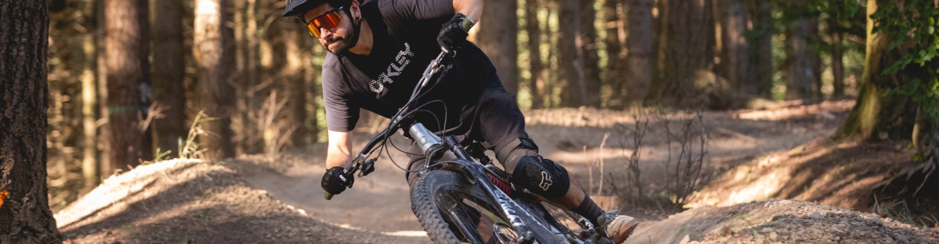 A mountain biker rides on a forest trail, surrounded by trees and a dirt path.