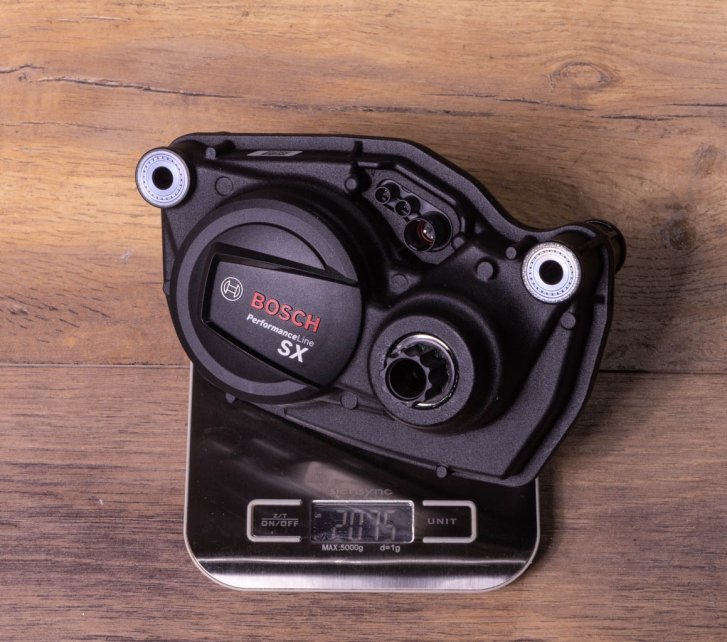 A Bosch SX e-bike drive on a kitchen scale on a dark wooden floor. Display shows 2,075 grams
