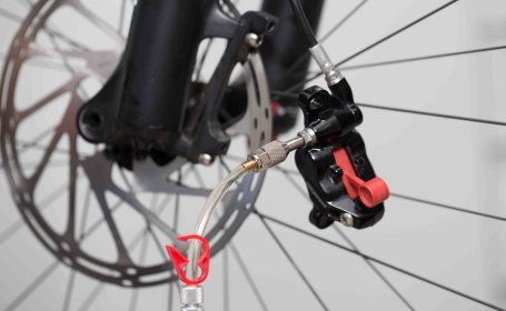 Ein losgeschraubter Scheibenbremssattel hängt lose an der Bremsleitung. An ihm ist ein Adapter zur Entlüftung angeschlossen. Im Hintergrund sind Fahrradspeichen und eine Bremsscheibe erkennbar.