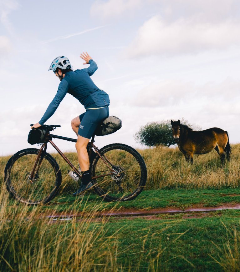 Ciclista de bikepacking Endura en bicicleta gravel saluda a un poni en un paisaje rural de colinas cubiertas de hierba durante una ruta fuera de carretera.