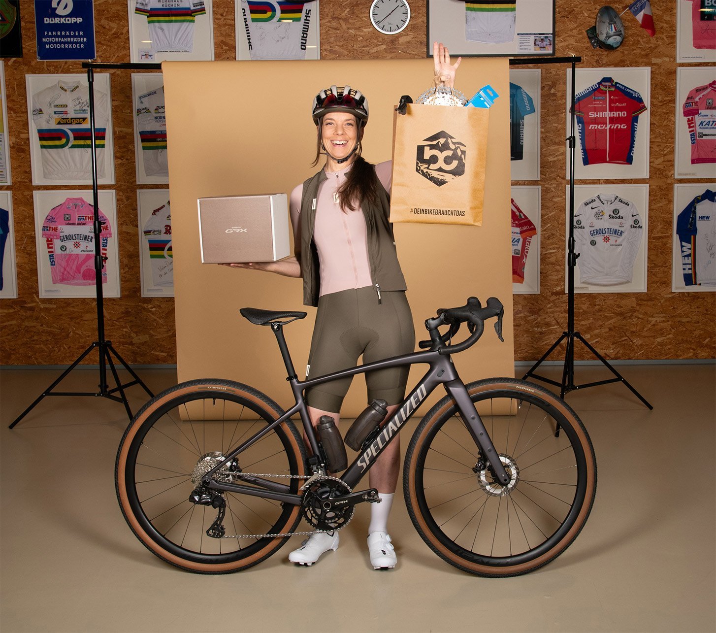Anke Eberhardt steht in Radbekleidung hinter einem Specialized Gravelbike und hält Shimano GRX Box und bc-Tasche in die Kamera.