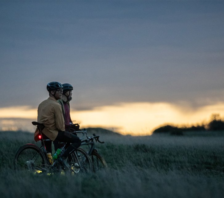 Dos ciclistas con cascos están con sus bicicletas en un campo al atardecer. Sus bicicletas tienen luces, el cielo es azul oscuro con un horizonte naranja y los árboles son visibles al fondo.