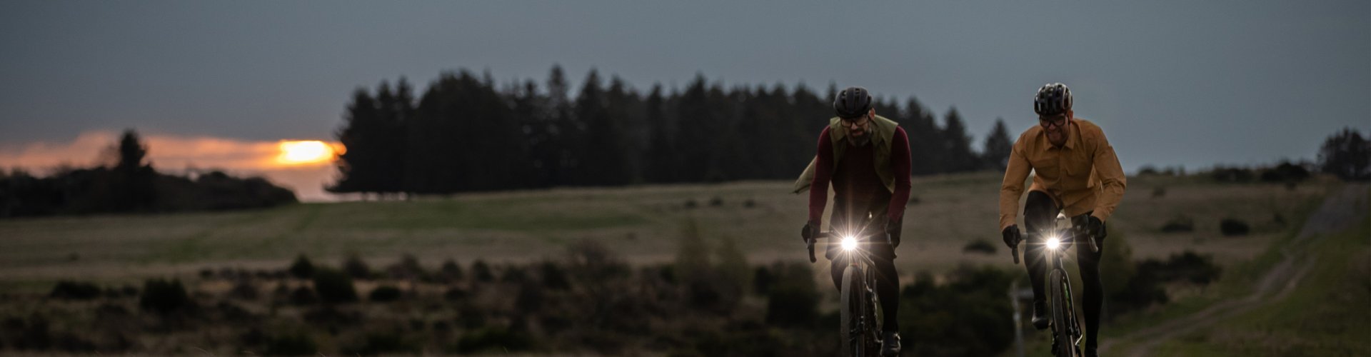 Dos personas montan bicicletas a través de un campo al atardecer. Sus bicicletas tienen luces. El cielo está nublado, el horizonte naranja, con árboles al fondo.