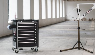 Werkzeugwagen und Montageständer in einer leeren Industriehalle mit großen Fenstern.