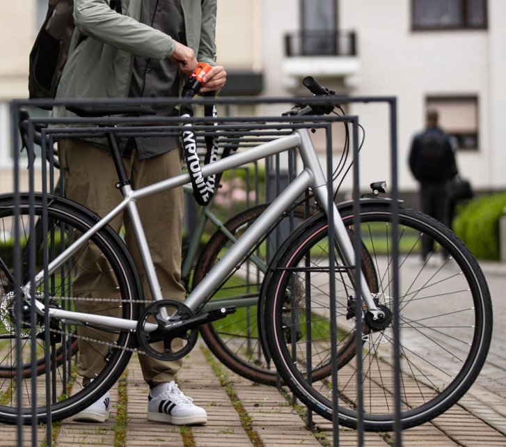 Vélo garé dans un support métallique devant un immeuble ; une personne le sécurise avec un antivol Kryptonite orange fixé au cadre.