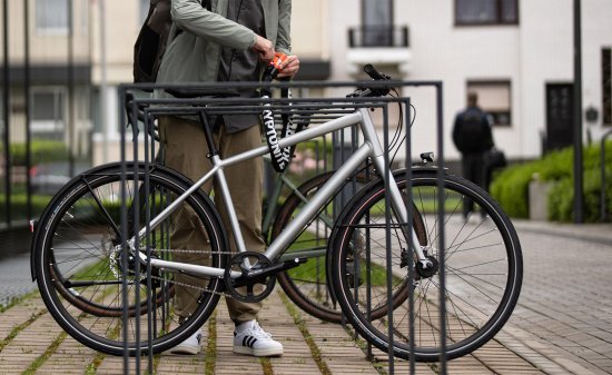 Vélo garé dans un support métallique devant un immeuble ; une personne le sécurise avec un antivol Kryptonite orange fixé au cadre.