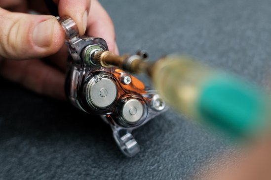 Una pinza de freno de 4 pistones de Trickstuff con carcasa transparente se está llenando con una jeringa. Se pueden ver claramente los pistones de acero y dos burbujas de aire.