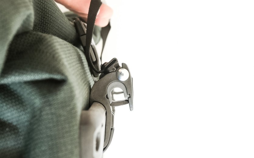 Nahaufnahme des Befestigungssystems einer grünen Ortlieb Back-Roller Urban Fahrradtasche. Eine Hand hält den schwarzen Verschlussgurt, während das Haken-System für die Montage am Gepäckträger sichtbar ist.