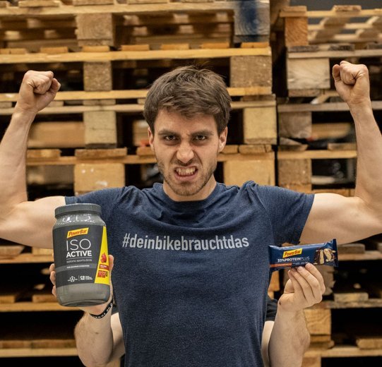 A man flexes his muscles. Behind him, another man is holding various nutritional supplements up to the camera.