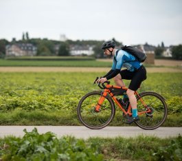 Homme portant un casque et un sac à dos roule de manière sportive sur un vélo de route le long d’un chemin asphalté à travers des champs verts ; maisons et arbres au loin.