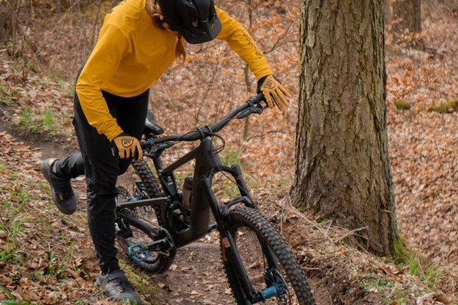 Una mujer con maillot ocre y casco oscuro de MTB se sube a una bicicleta de montaña oscura.