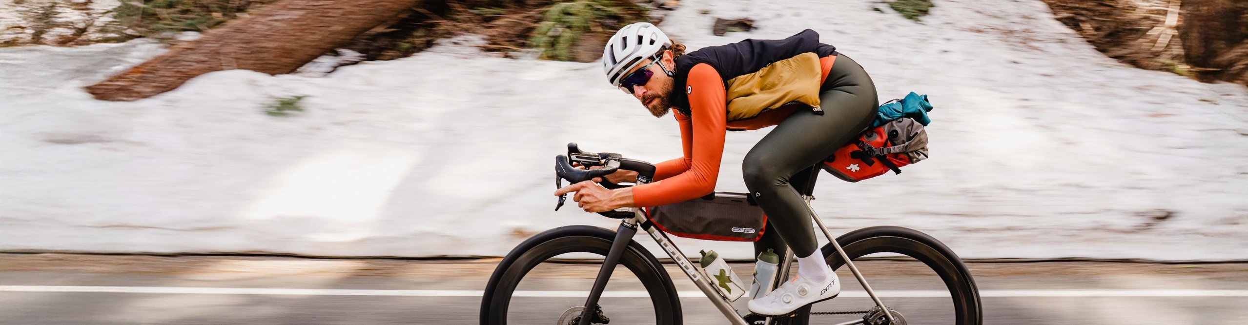 Rennradfahrer in Assos-Bekleidung fährt dynamisch durch verschneite Berglandschaft mit voll bepacktem Bikepacking-Setup.