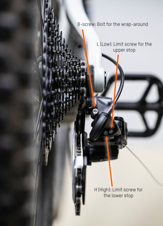 The MTB rear derailleur can be seen from behind. The limit screws H and L, as well as the B-bolt are marked with orange arrows.