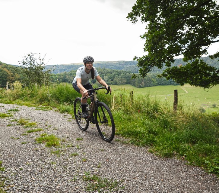 Susi from bc's Social Media Team rides a bc original Flint gravel bike over a gravel track surrounded by green, hilly countryside. 