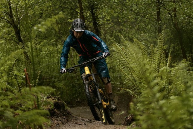 A mountain biker in blue rain gear and a black helmet rides along a narrow, muddy trail through a dense, green forest.