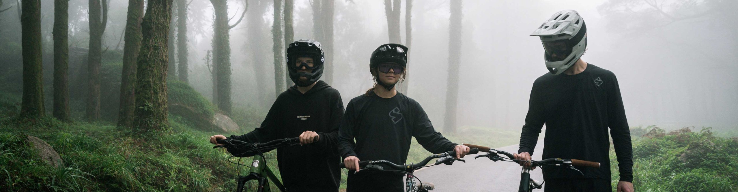 Tres ciclistas de montaña con cascos Sweet Protection empujan sus bicicletas por una carretera mojada en un bosque con niebla.