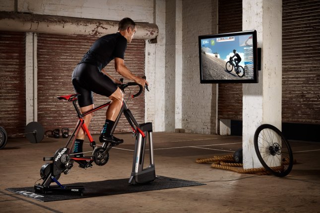 A man trains on a direct-drive smart trainer in front of a screen inside a gym with whitewashed brick walls and a steel roll-up door.
