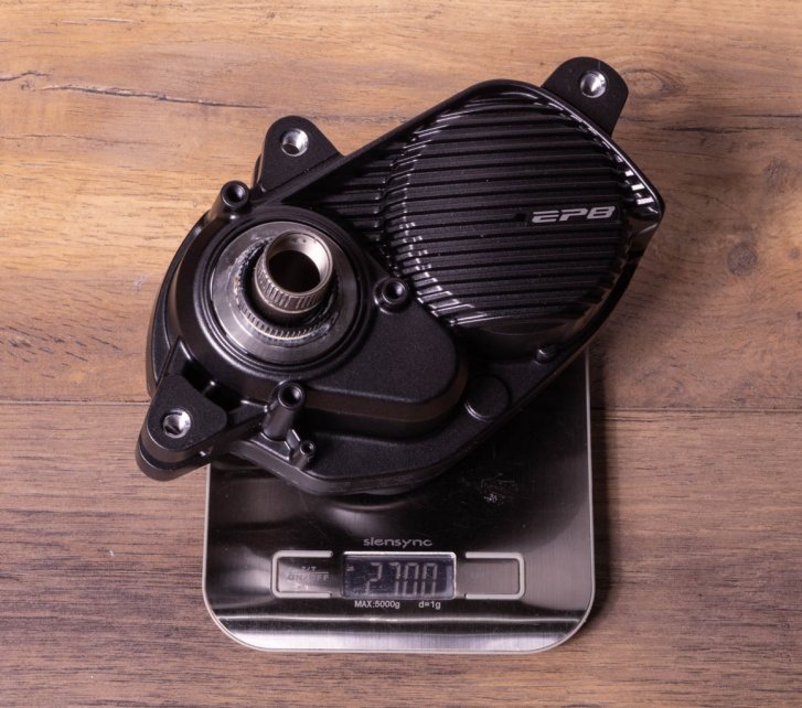 A Shimano EP8 e-bike drive on a kitchen scale on a dark wooden floor. Display shows 2,700 grams