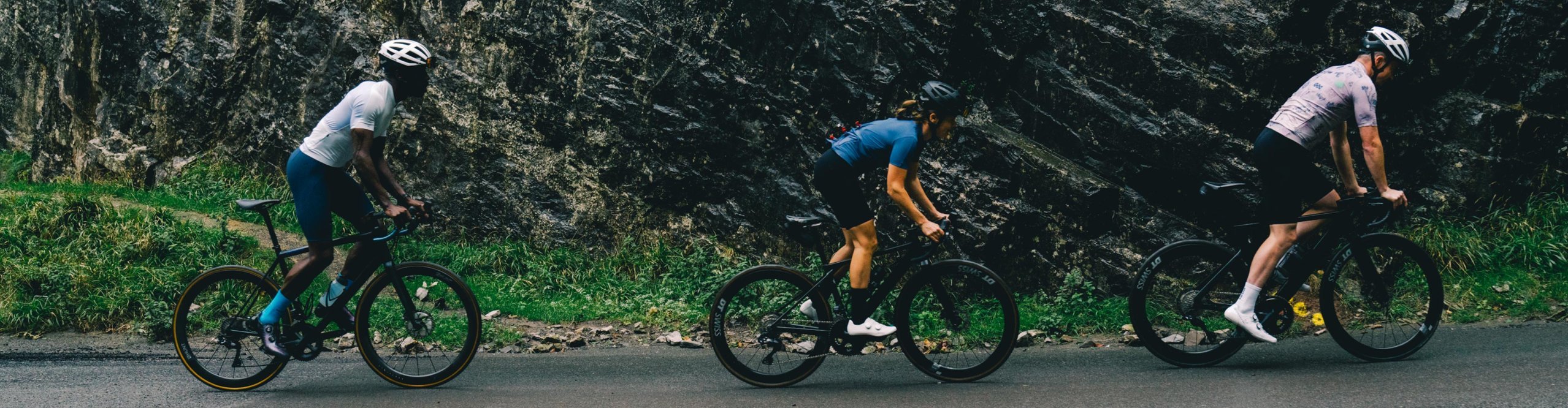 Tres ciclistas de carretera con ropa de ciclismo Endura pedaleando por una carretera de montaña junto a una pared rocosa