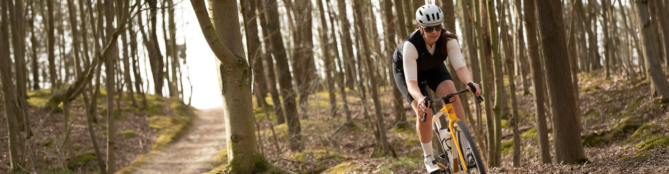 Gravelbikerin fährt mit einem Gravelbike auf einem schmalen Waldtrail durch einen lichten Laubwald. Sie trägt Helm, Sonnenbrille und leichte Fahrradbekleidung für sportliche Fahrten auf Schotter und Naturwegen.