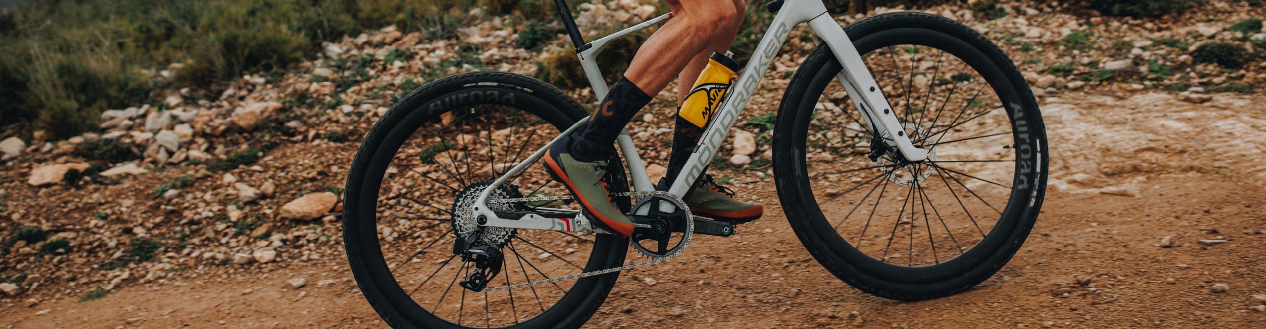Ciclista de gravel con casco Mavic y ropa de ciclismo Mavic sobre una bicicleta gravel blanca en un camino pedregoso bajo cielo nublado