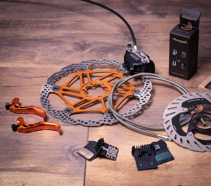 Algunas piezas de mejora para frenos de disco sobre una superficie de madera: discos, latiguillo reforzado, pastillas con aletas y dos palancas anodizadas en naranja.