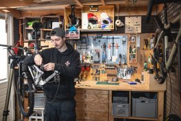 Ein junger, schlanker Mann mit herumgedrehtem Käppie, dunklem Schnauzbart und schwarzem Hoodie arbeitet in einer aufgeräumten Fahrradwerkstatt.