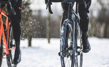Pictured are two bikers during a gravel tour in the snow. The focus of the photo is on the bikes. Both cyclists are wearing shoe covers. 