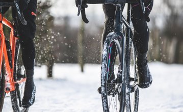 La photo montre deux cyclistes lors d'une sortie gravel dans la neige. L'image se concentre sur les vélos. Les deux cyclistes portent des surchaussures. 