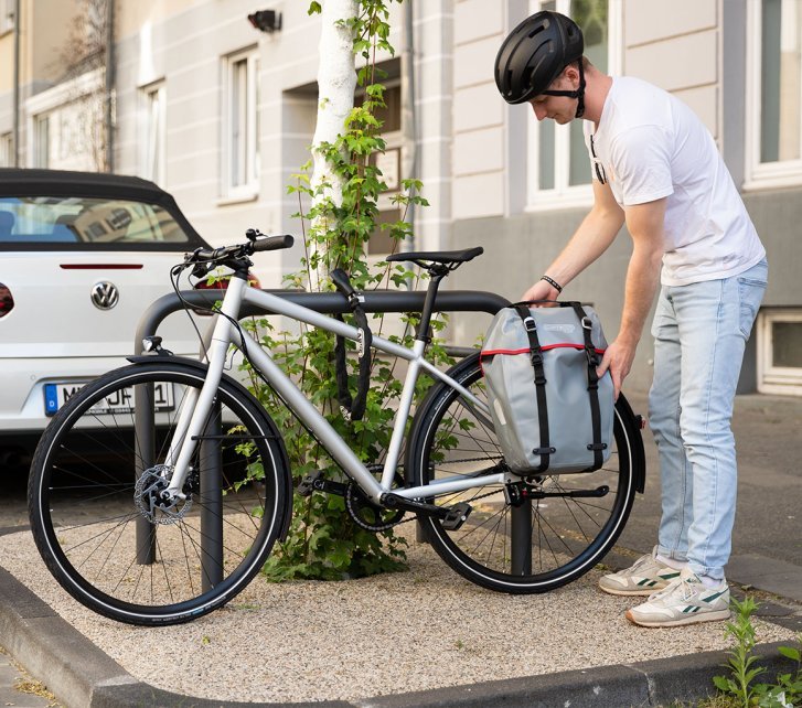 A person locks a bicycle with a pannier bag to a bike rack in an urban setting, next to a parked car and a building with ivy.