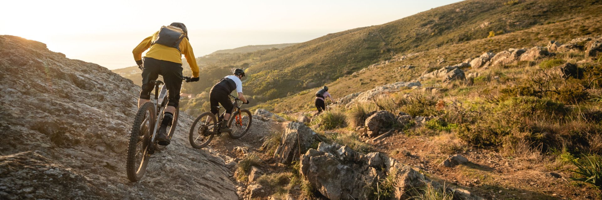 Drei E-Mountainbiker befahren einen felsigen Trail im Gegenlicht. Wir sehen die drei von hinten, die Abendsonne taucht alles in ein goldenes Licht.