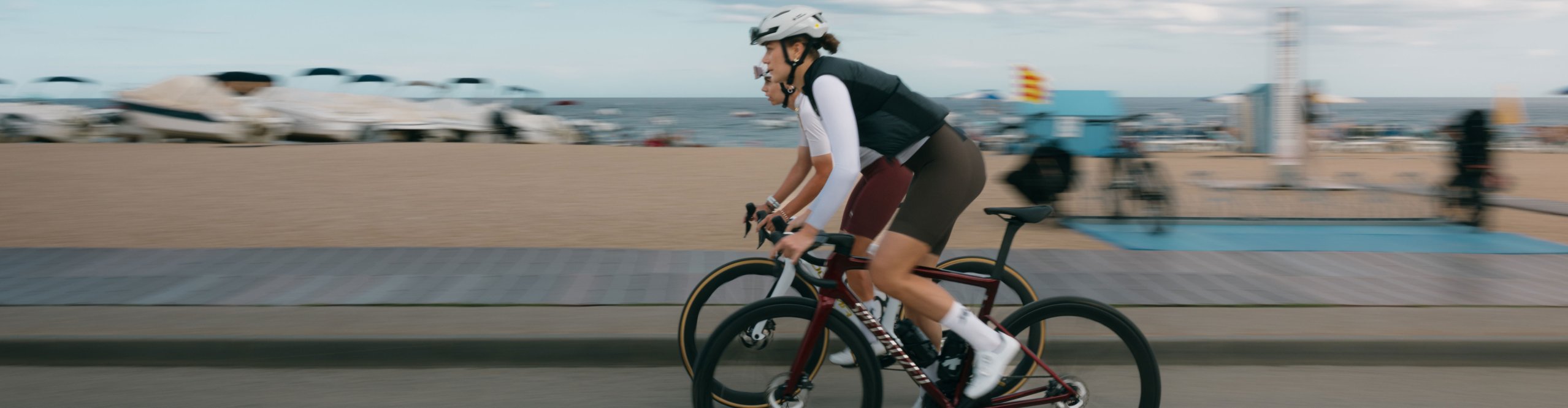 Dos ciclistas de carretera con outfit de Assos ruedan por un paseo marítimo, con playa y mar de fondo.