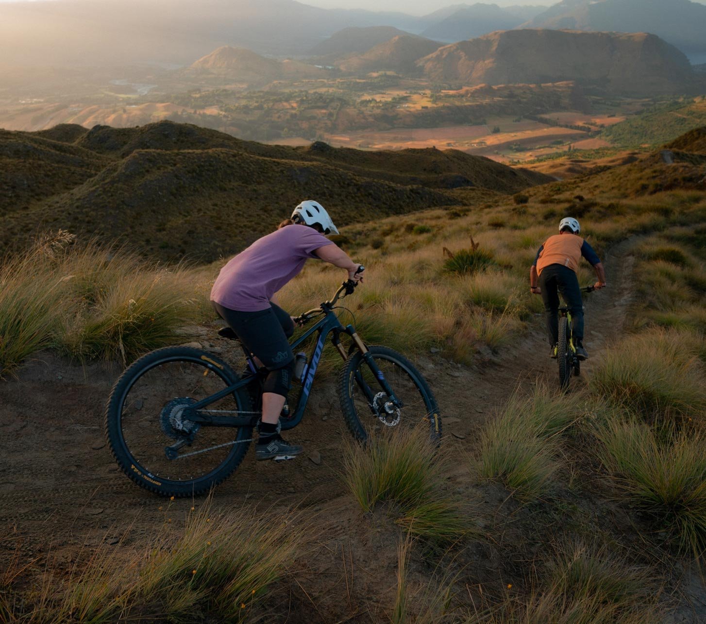 Two mountain bikers ride a trail at golden hour, featuring a Pivot Firebird enduro mountain bike in the foreground.