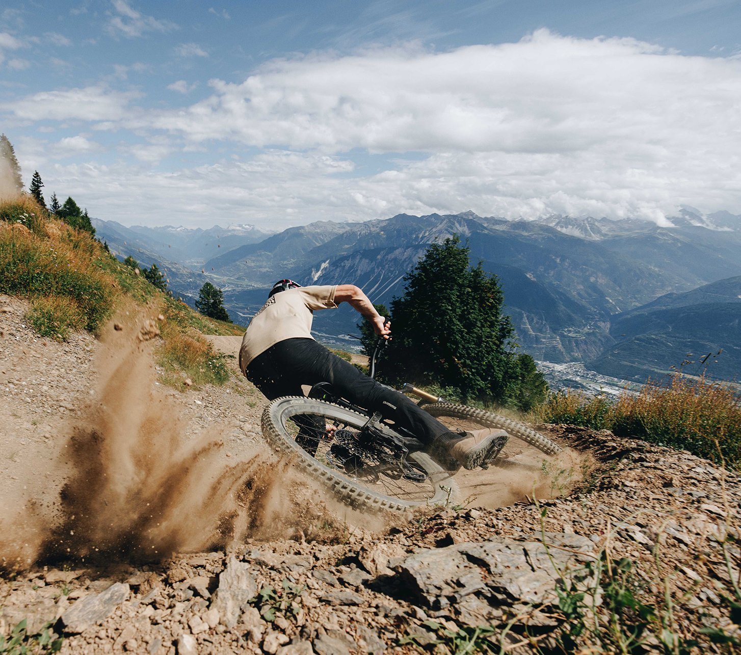 Mountainbiker driftet mit viel Speed durch eine staubige Kurve auf einem alpinen Trail, spektakuläre Bergkulisse im Hintergrund