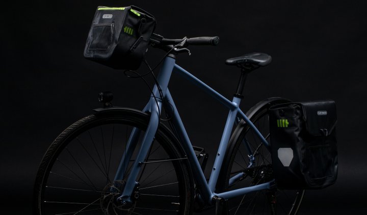 Una bicicleta azúl con una bolsa de manillar ORTLIEB negra y una alforja trasera, ambas con detalles verdes. La bolsa de manillar tiene un diseño iluminado. El fondo es oscuro.