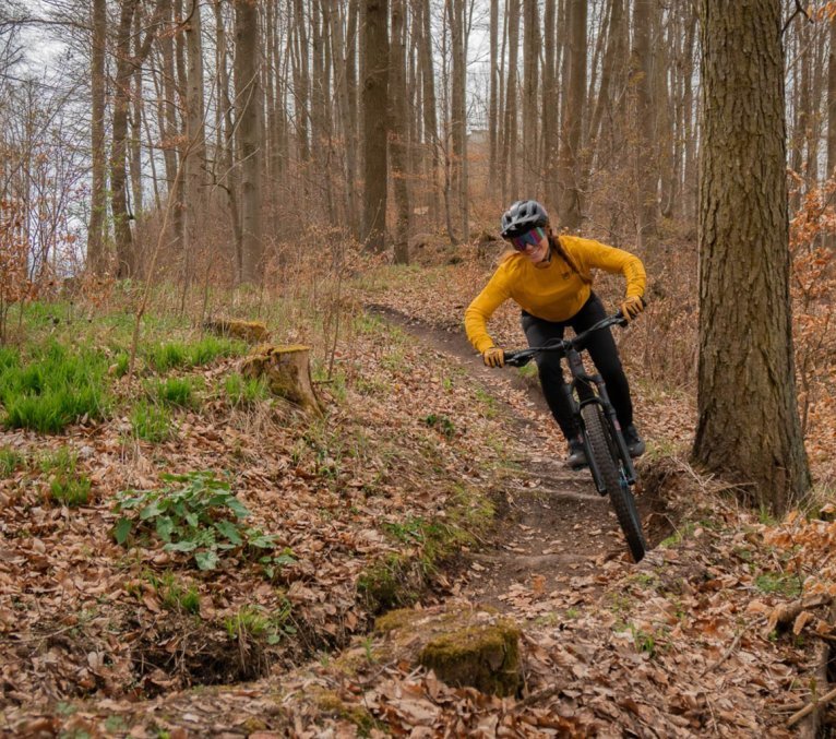 Mujer con maillot amarillo de MTB, casco negro y gafas de colores sonríe y señala su maillot mientras está sentada en una bicicleta de montaña en un bosque otoñal.
