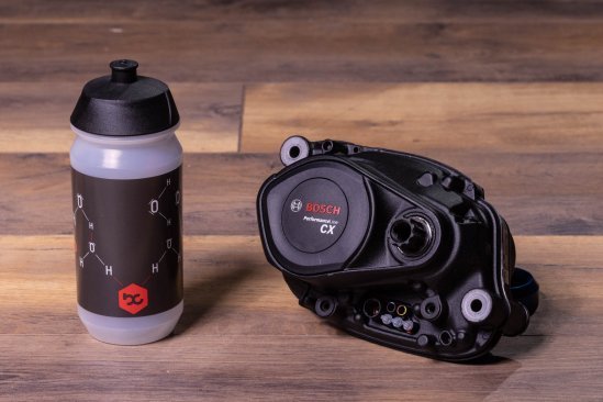 A Bosch Performance CX e-bike drive lies on a dark wooden floor. Next to it is a bike water bottle