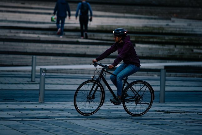 Un ciclista con luces en su bicicleta y casco pedalea en una plaza pavimentada de la ciudad por la noche, mientras dos personas caminan al fondo.