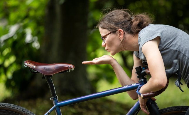 A person in a gray T-shirt leans over a bicycle and kisses the brown saddle. The bike has a blue frame and is positioned in a green, wooded environment.