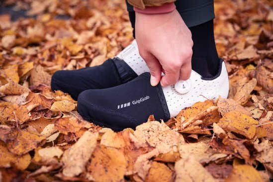 Dos pies con zapatillas gravel blancas y toe caps negras; una mano ajusta la cubierta.