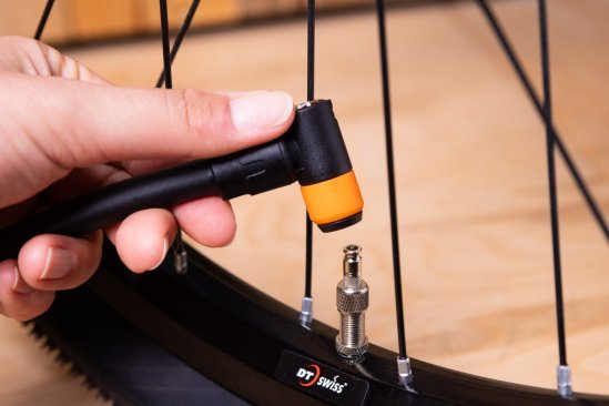 Close-up: A hand holds a black Clik Valve pump head with an orange ring next to a mounted Clik Valve.