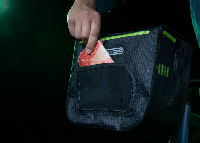 Eine Hand steckt ein rotes Tuch in die Seitentasche einer schwarzen Fahrradtasche mit grünen Akzenten. Die Tasche hat ein beleuchtetes Design. Der Hintergrund ist dunkel.