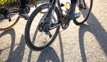 Two road cyclists are riding in winter weather. The focus of the picture is on the drivetrain of the cyclist on the right. 