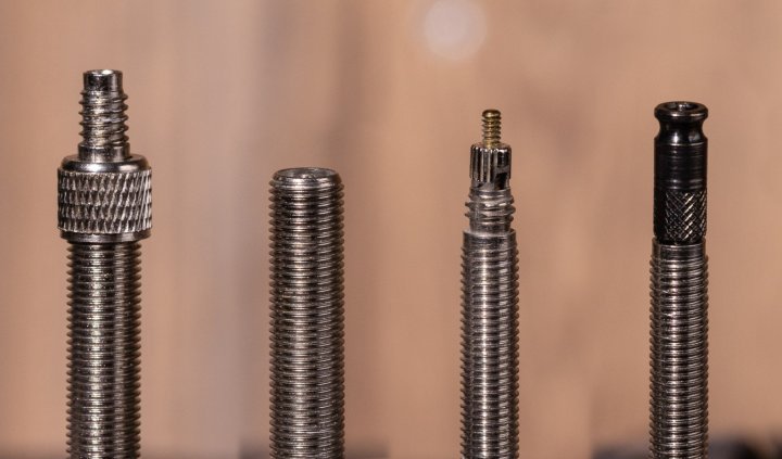 Makro-Foto: Ein Dunlop-Ventil, ein Autoventil, ein Rennrad-Ventil und ein Schwalbe Clik-Valve nebeneinander vor einer unscharfen braunen Holzoberfläche