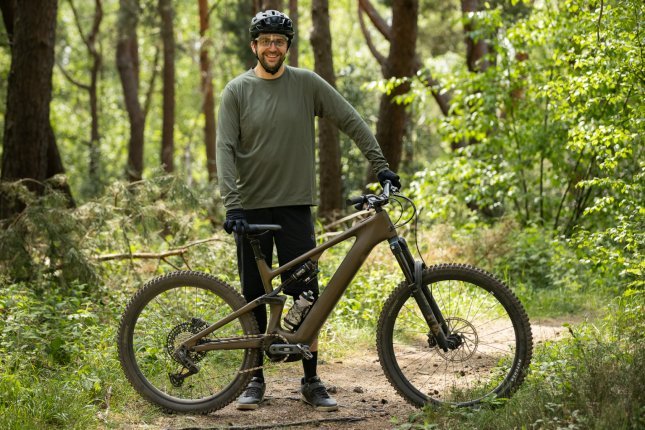 Berno steht im schwarz-oliven MTB-Outfit mit schwarzem Helm hinter dem Specialized Turbo Levo 4 im Wald und grinst in die KAmera.