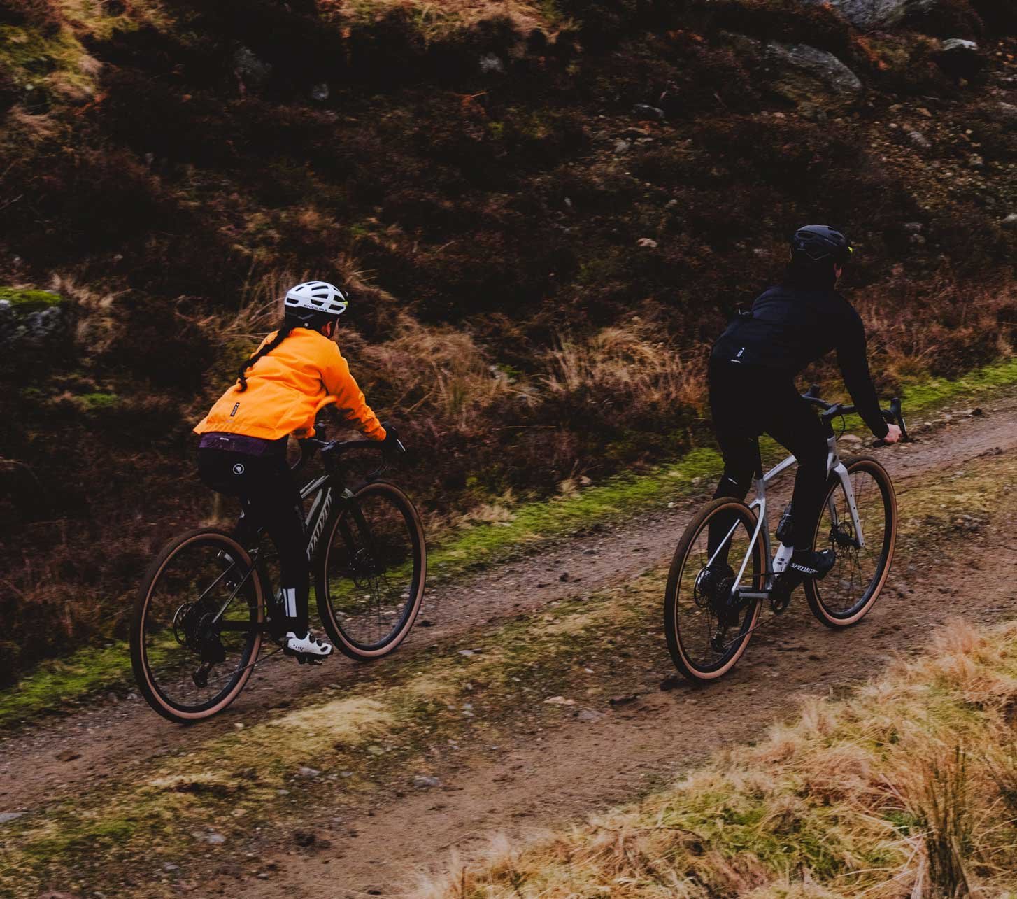 Deux cyclistes gravel portant des vestes Endura roulent sur un chemin humide à travers un paysage vallonné.