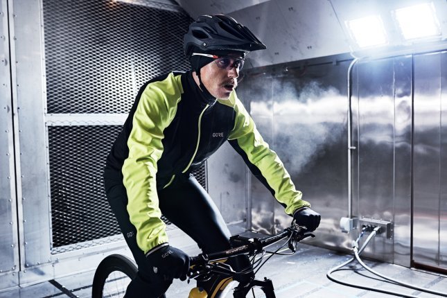 A cyclist wearing a black and yellow jacket and a black helmet rides inside a wind tunnel under artificial lighting.
