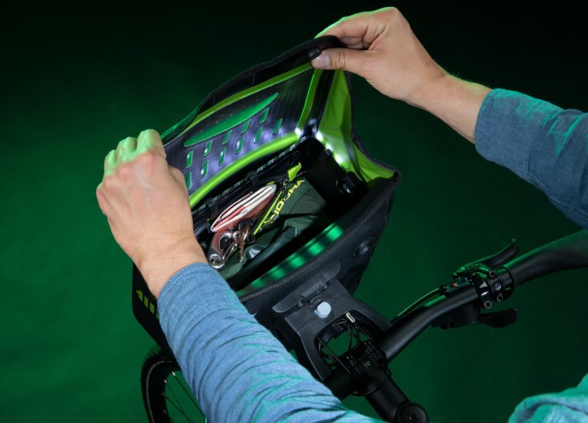 Zwei Hände öffnen eine schwarze Fahrradtasche mit grünen Akzenten am Lenker. Die Tasche hat ein beleuchtetes, perforiertes Design und enthält Gegenstände wie Schlüssel. Der Hintergrund ist dunkelgrün.